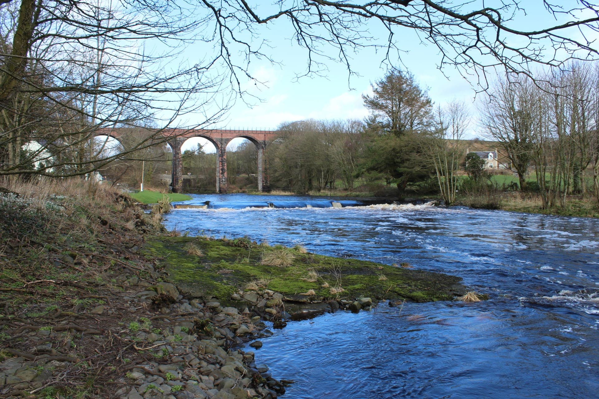 The Water of Luce at New Luce on a bright day, with the railway viaduct beyond